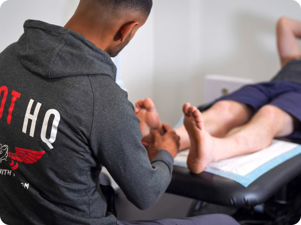 Podiatrist in a gray hoodie with 'FOOT HQ' on the back examining a person's foot.